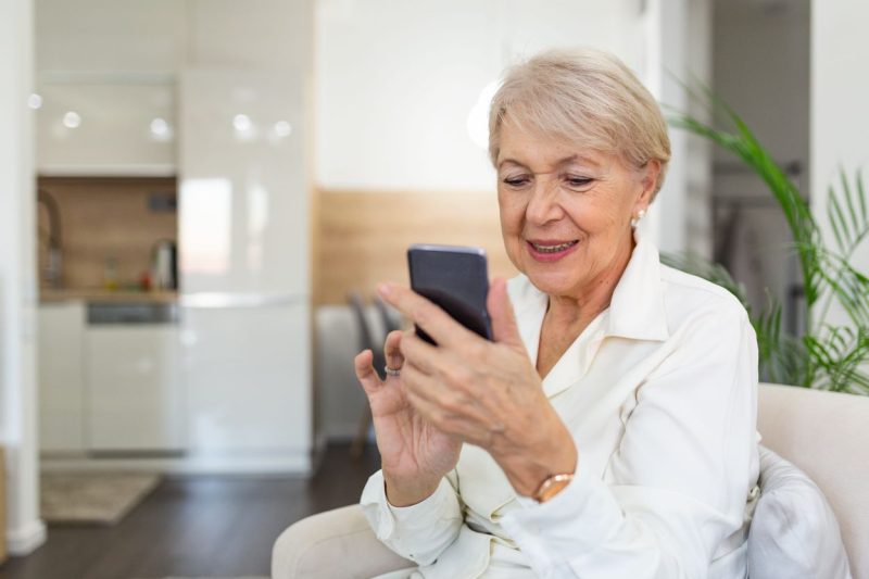 A senior woman smiles while using a smartphone at home, representing modern mobile-friendly online learning.